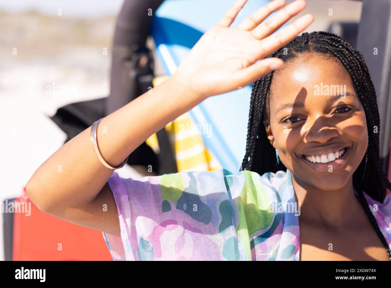 Jeune femme afro-américaine sourit brillamment à l'extérieur lors d'un Road trip. Son expression joyeuse et sa vague suggèrent une ambiance décontractée et ensoleillée de jour de plage. Banque D'Images
