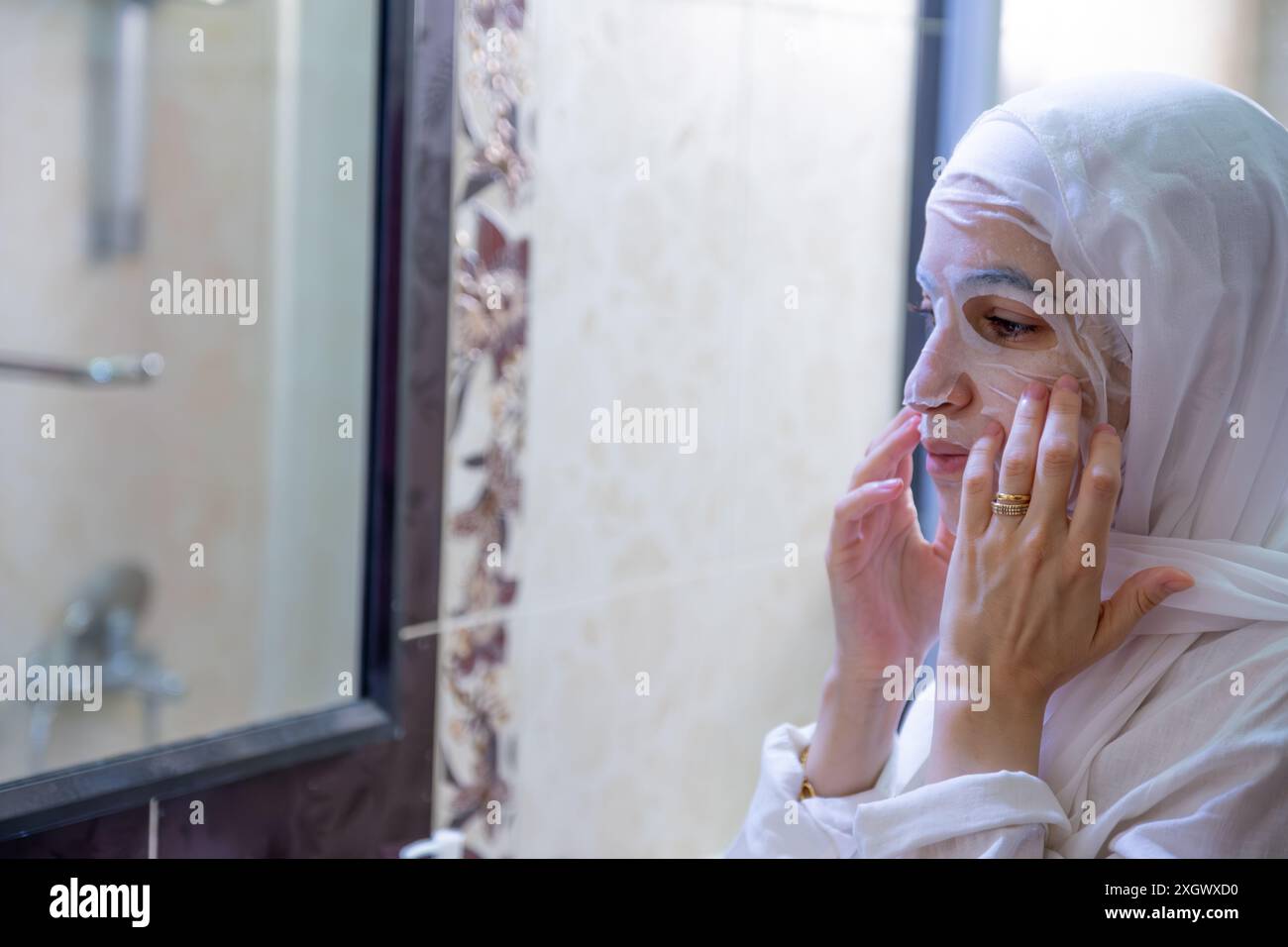 Femme musulmane arabe portant le hijab appliquant un masque facial dans le spa ou le bain regardant son visage et sourire Banque D'Images