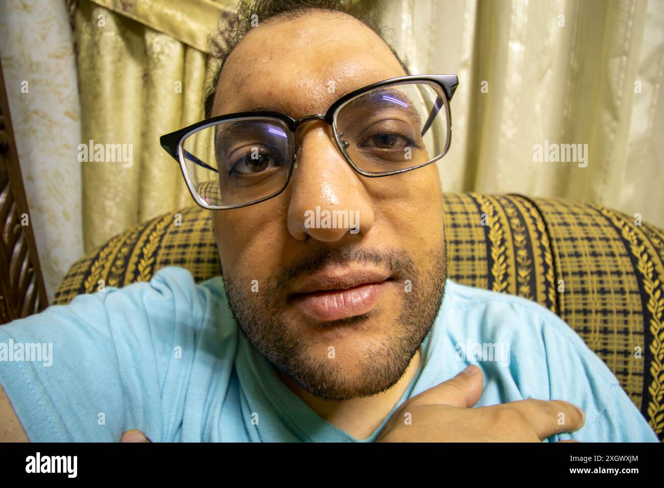 Un homme avec des lunettes prend un selfie intérieur détendu, mettant en valeur son style de vie décontracté et confortable. Son expression franche, sa barbe et son fond highli Banque D'Images
