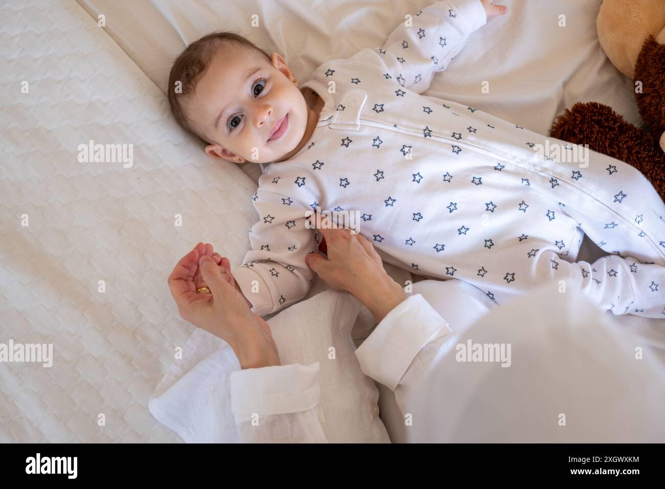 Bébé en pyjama à motifs étoilés couché sur un lit blanc avec un ours en peluche, assisté par un parent en hijab. Vue de dessus, gros plan, parfait pour la famille et les parents Banque D'Images