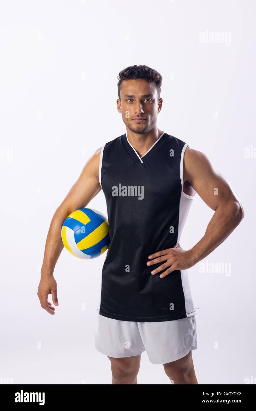 Athlétique jeune homme biracial pose avec un volley-ball sur un fond blanc. Sa position confiante suggère une préparation pour un match compétitif ou un entraînement Banque D'Images