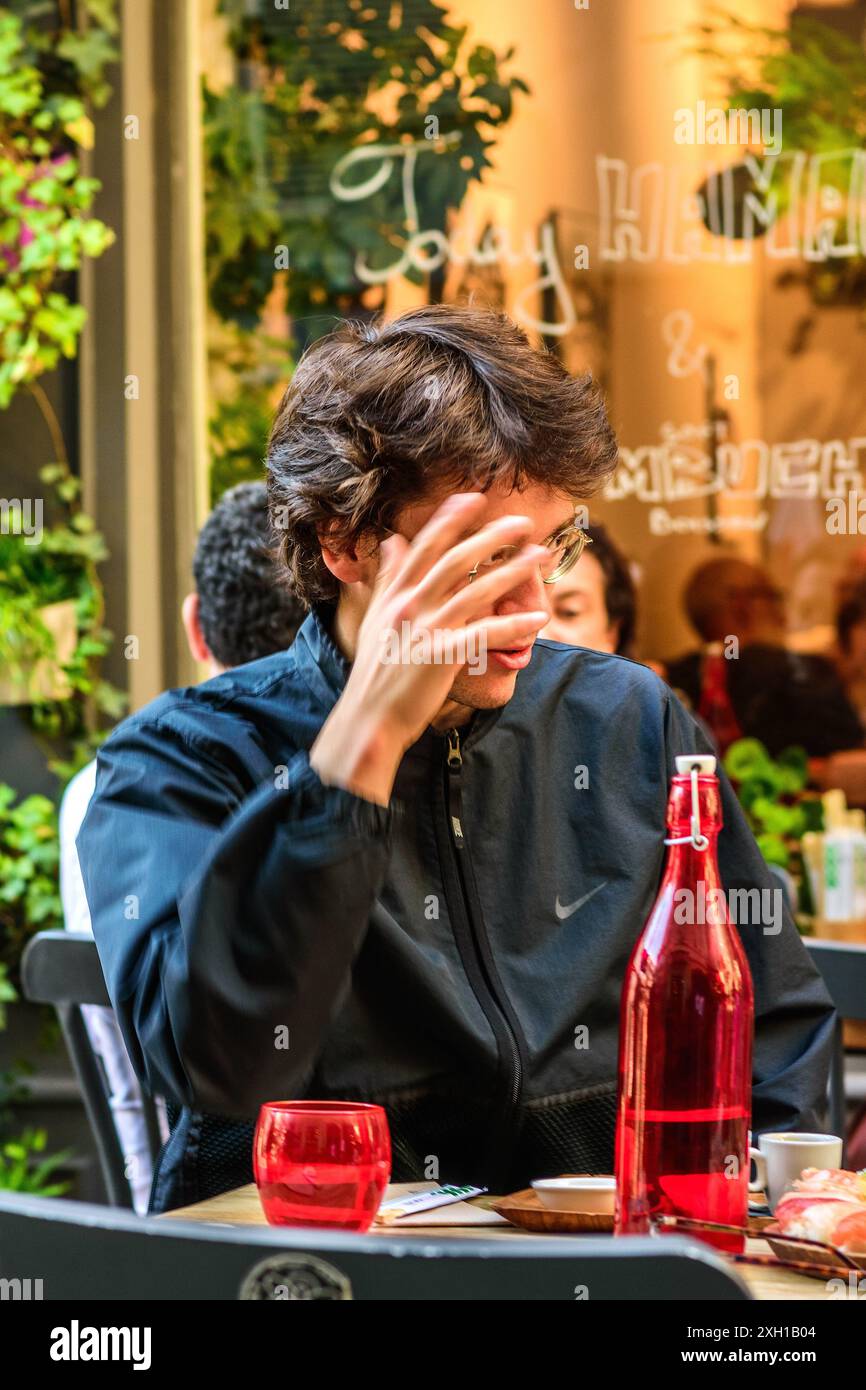Jeune homme à la table de café en plein air gesticulant avec la main - Paris 75004, France. Banque D'Images