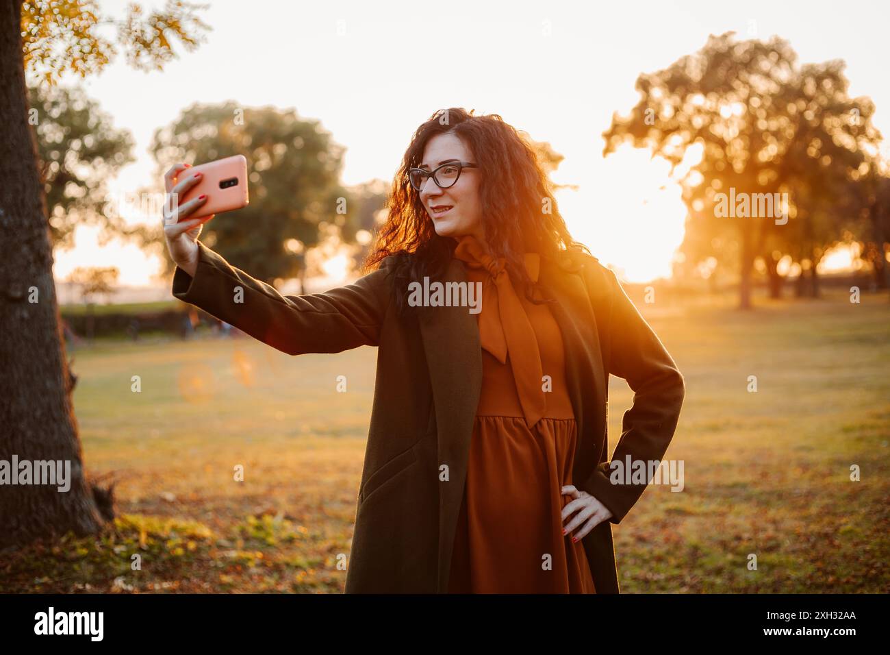 Une jeune femme aux cheveux longs et bouclés et aux lunettes prend un selfie avec son téléphone dans un parc. Elle porte un manteau marron et une robe orange. Le paramètre Banque D'Images