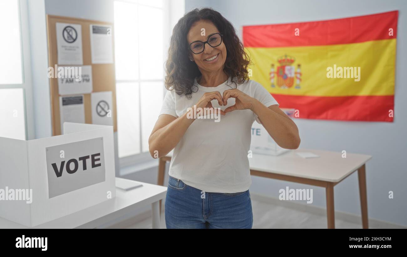 Femme hispanique faisant symbole de coeur avec des mains dans une salle électorale à l'intérieur avec une bannière de drapeau espagnol en arrière-plan pendant les élections. Banque D'Images