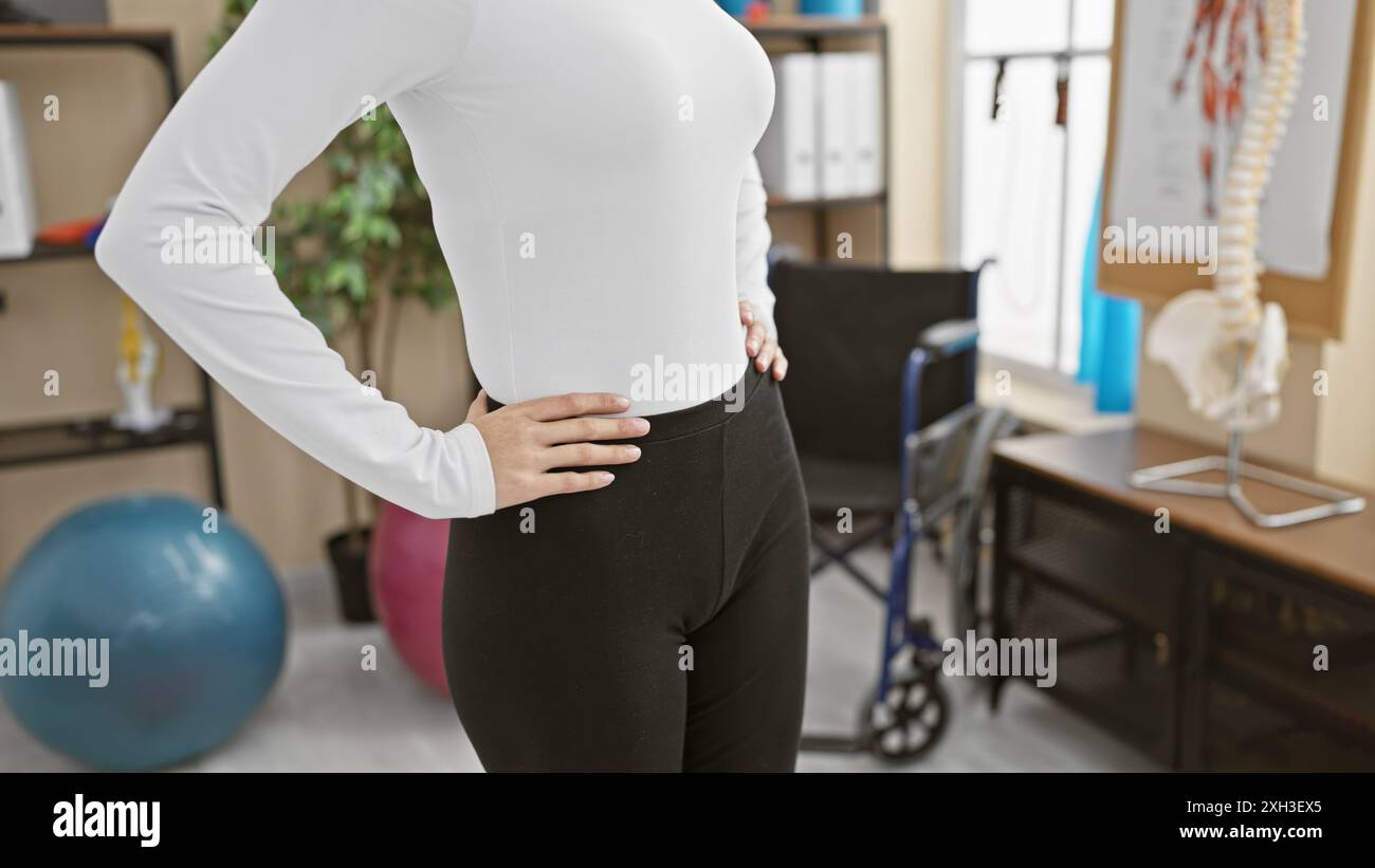 Femme dans une salle de physiothérapie tient sa hanche, portant une tenue de gymnastique, entourée de ballons d'exercice et de matériel médical. Banque D'Images