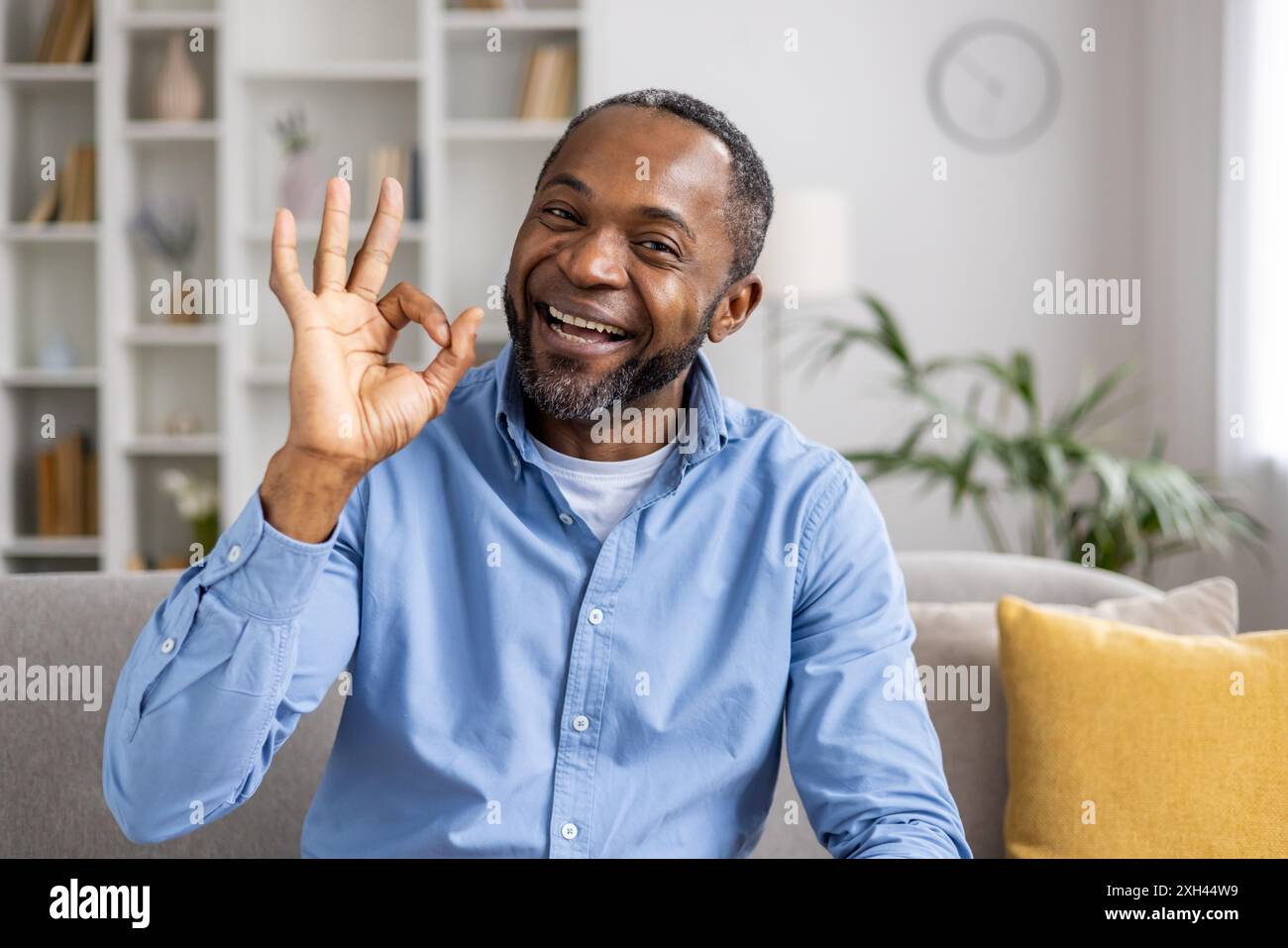Homme souriant affichant le geste OK, exprimant la positivité et le bonheur dans le salon lumineux. Cadre décontracté avec canapé confortable, plantes décoratives et oreillers moelleux. Banque D'Images