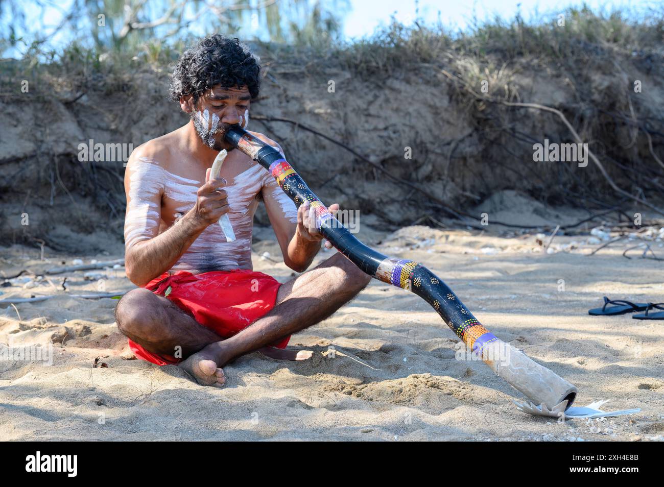 Un aborigène vêtu de vêtements traditionnels et de peinture corporelle assis sur la plage de k'gari , jouant du didgeridoo Banque D'Images