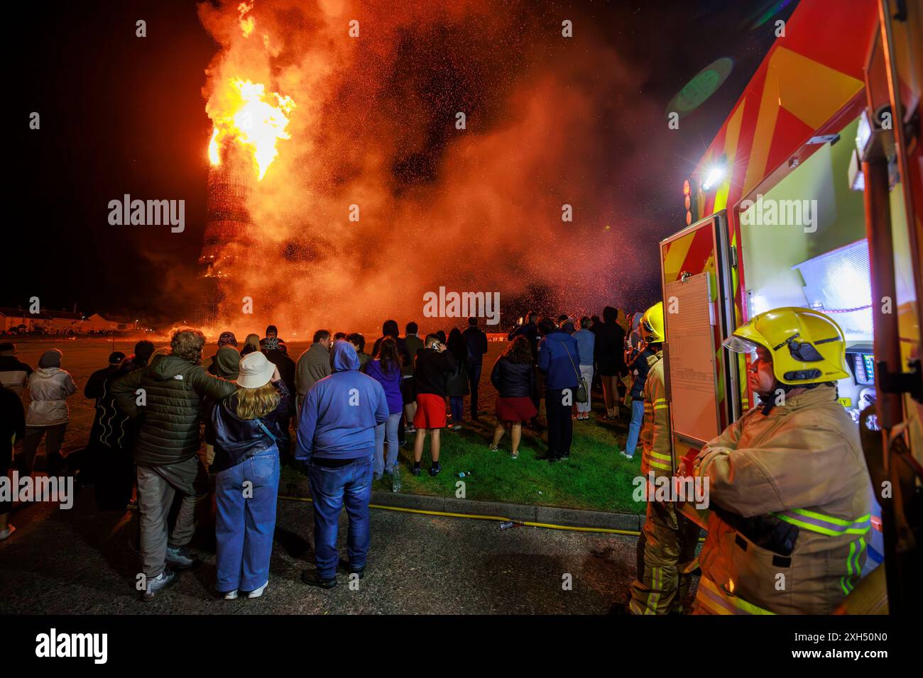Des membres du public et des pompiers regardent le bûcher en feu de camp de la onzième nuit de Craigyhill à Larne. L'incendie des feux de joie loyalistes fait partie des traditionnelles douzième commémorations marquant l'anniversaire de la victoire du roi protestant William sur le roi catholique James à la bataille de la Boyne en 1690. Date de la photo : vendredi 12 juillet 2024. Banque D'Images