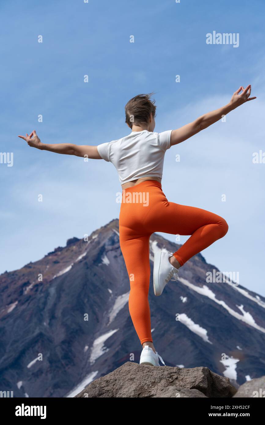 Vue arrière d'une femme pratiquant le yoga, debout sur le sommet d'une montagne rocheuse avec un volcan en arrière-plan. Femme galbée équilibre le corps sur une jambe. Professeur de yoga Banque D'Images