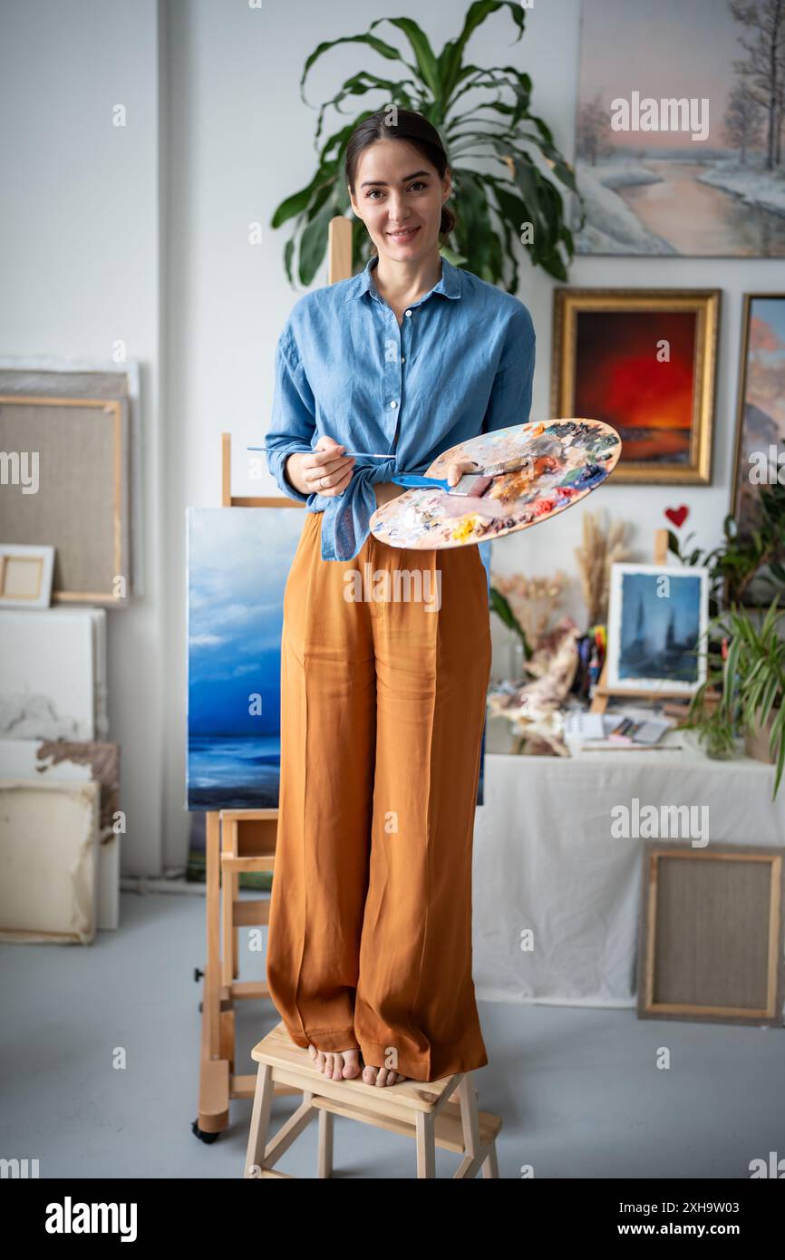 Portrait d'artiste féminine heureuse réussie debout sur une chaise avec palette de peintures à l'huile, pinceaux Banque D'Images