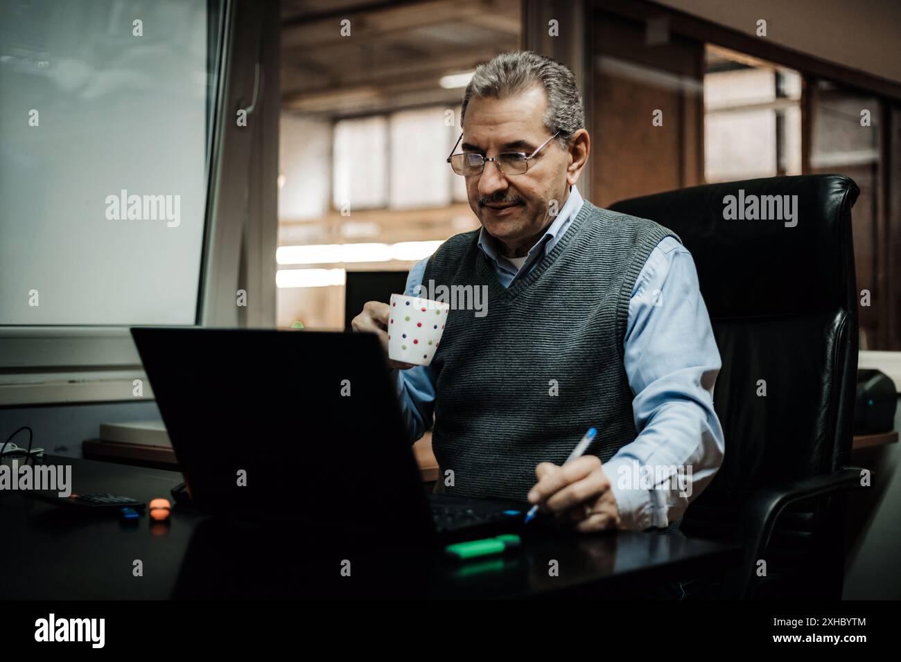 Un homme est assis à un bureau dans un bureau, travaillant sur un ordinateur portable et tenant une tasse à café. Il porte une chemise bleue et un gilet de pull gris. Banque D'Images