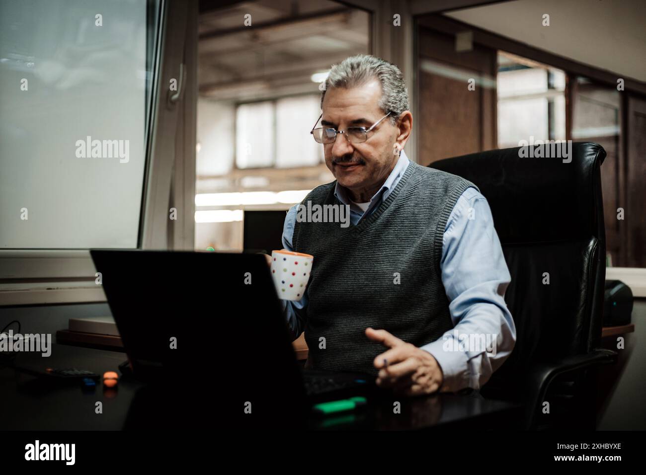 Un homme est assis dans une chaise de bureau noire, regardant son ordinateur portable, avec une tasse à café dans sa main droite. Il est vêtu d'une chemise bleue et d'un gilet gris. Banque D'Images
