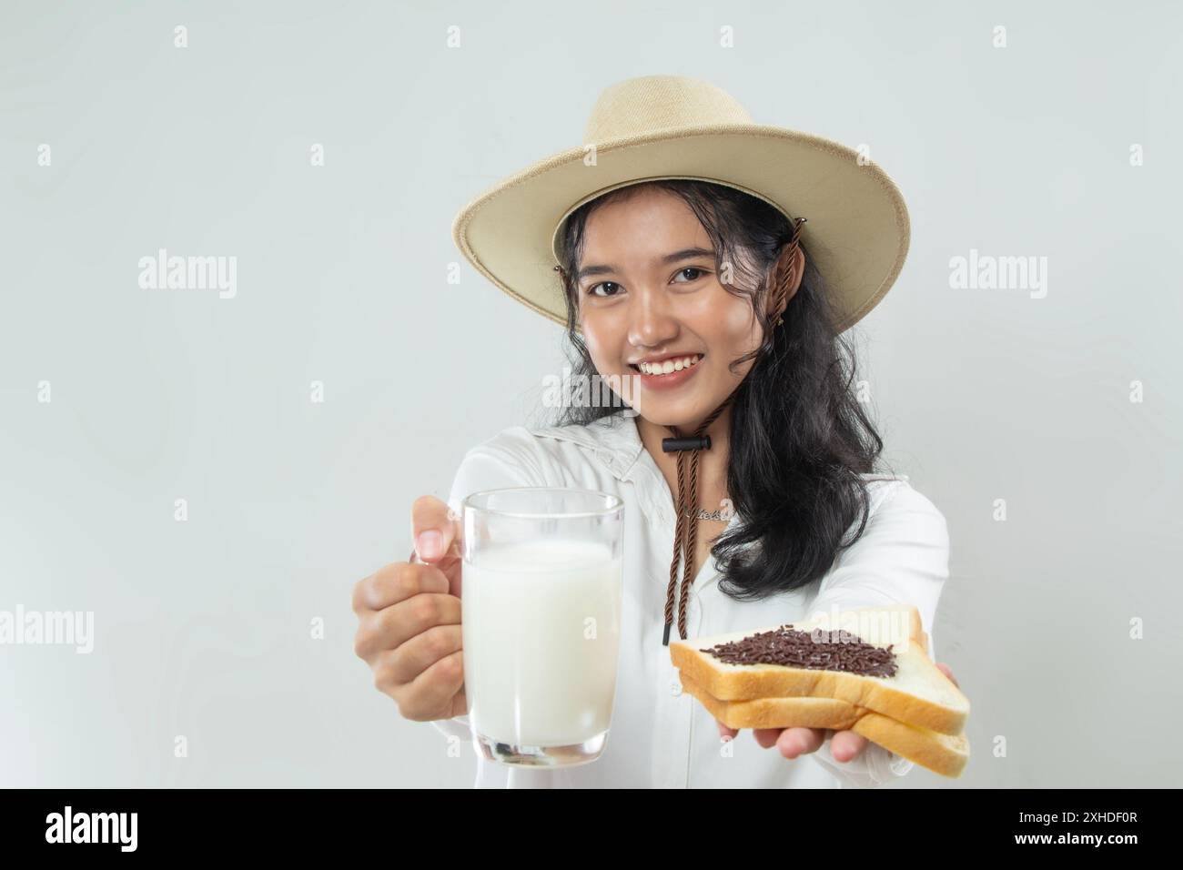 Belle jeune femme asiatique en chapeau de cow-boy distribuant du lait, du pain blanc saupoudré de chocolat au lait tartiné à la caméra avec sourire heureux, petit déjeuner co Banque D'Images