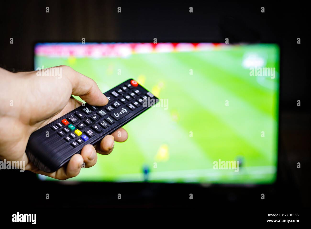 La main d'une personne tient une télécommande devant un écran de télévision montrant un match de football flou. Banque D'Images