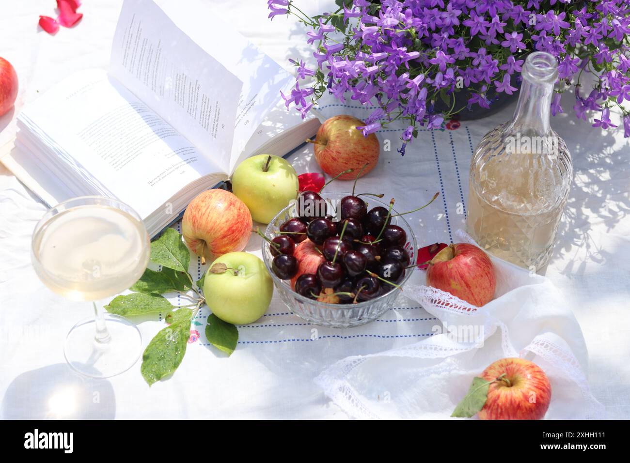 Nature morte avec un livre ouvert, cerise, pommes, verres à vin et bouquet de fleurs Campanula sur une table de jardin. Image lumineuse de table de pique-nique. Banque D'Images