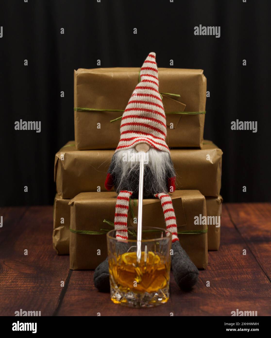 Un gnome de Noël assis sur un cadeau, buvant du whisky à travers une paille. Fond noir, plancher en bois, cadeaux enveloppés dans du papier recyclé Banque D'Images