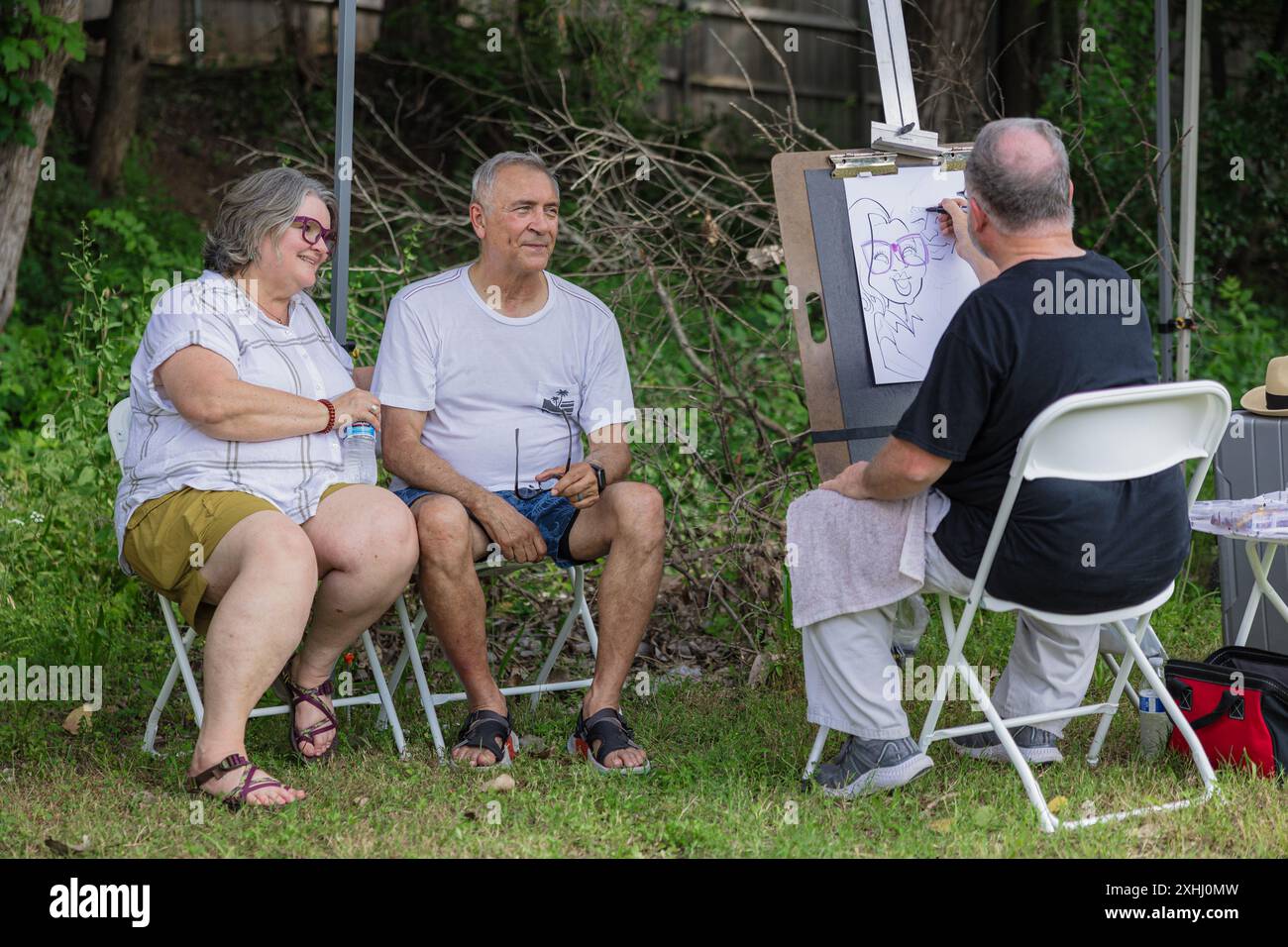 Un couple de personnes âgées aime un caricaturiste dessinant leur portrait à l'extérieur dans un cadre de parc Banque D'Images