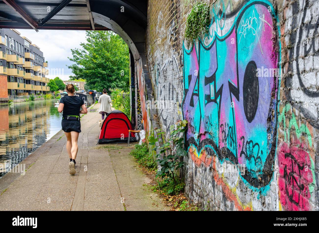 Une dame court le long d'un sentier le long du bras de Paddington du Grand Union canal devant une tente appartenant à un sans-abri à Londres, au Royaume-Uni Banque D'Images