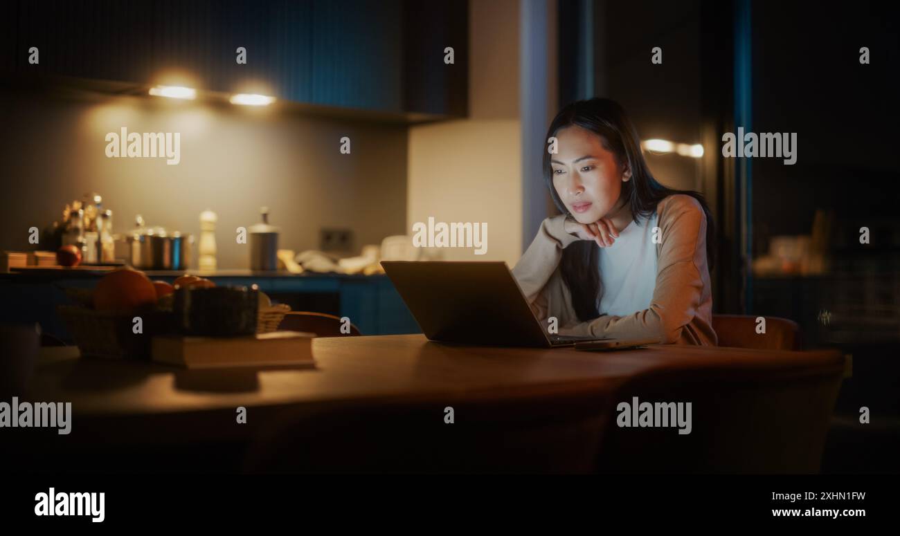 Belle femme asiatique authentique assise à une table dans une cuisine confortable et en utilisant un ordinateur portable à la maison la nuit. Femmes souriant et discutant sur Internet et les médias sociaux, faire de la recherche Banque D'Images