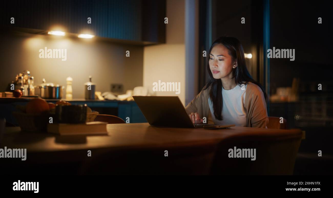 Belle femme asiatique authentique assise à une table dans Cozy Kitchen et à l'aide d'un ordinateur portable à la maison la nuit. Femmes souriant et discutant sur Internet et les médias sociaux, faisant de la recherche scientifique. Banque D'Images