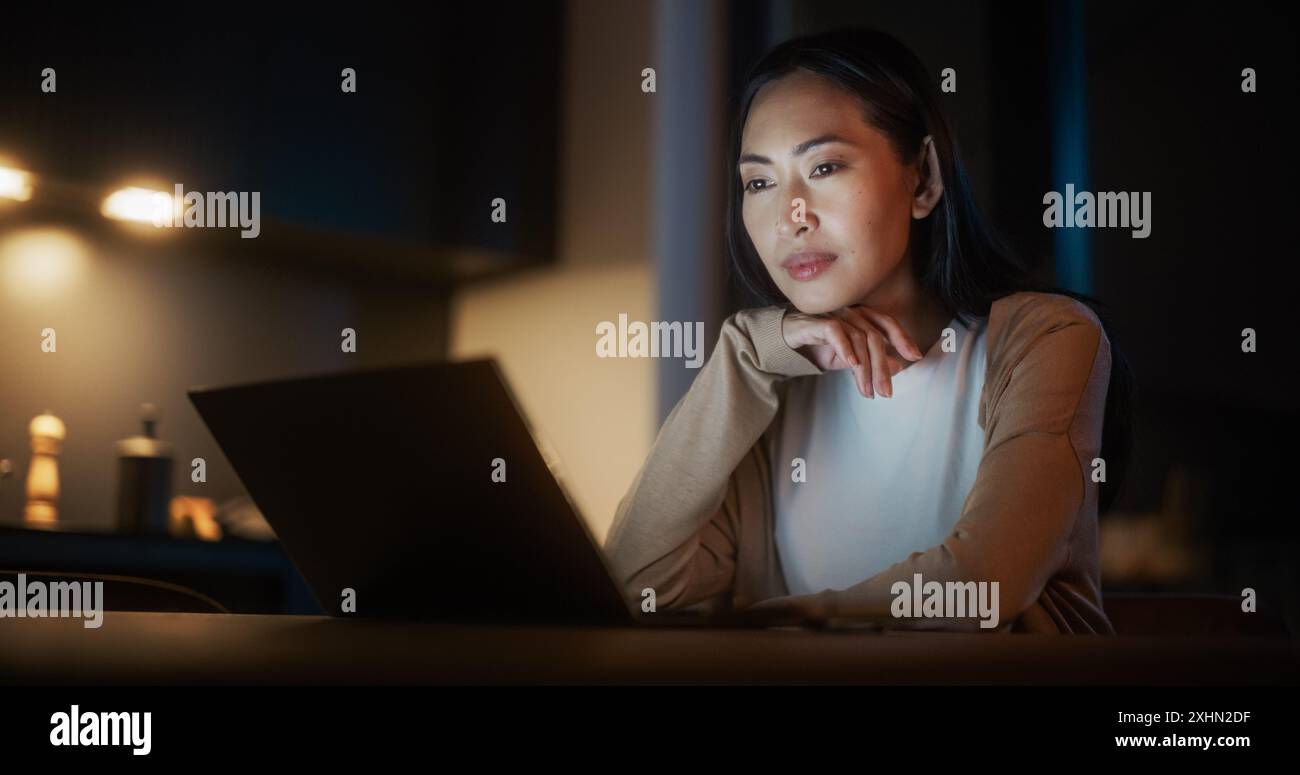 Belle femme asiatique authentique assise à une table dans une cuisine confortable et en utilisant un ordinateur portable à la maison la nuit. Femme souriant et discutant sur les médias sociaux, faisant des achats en ligne, vérifiant les nouvelles Banque D'Images