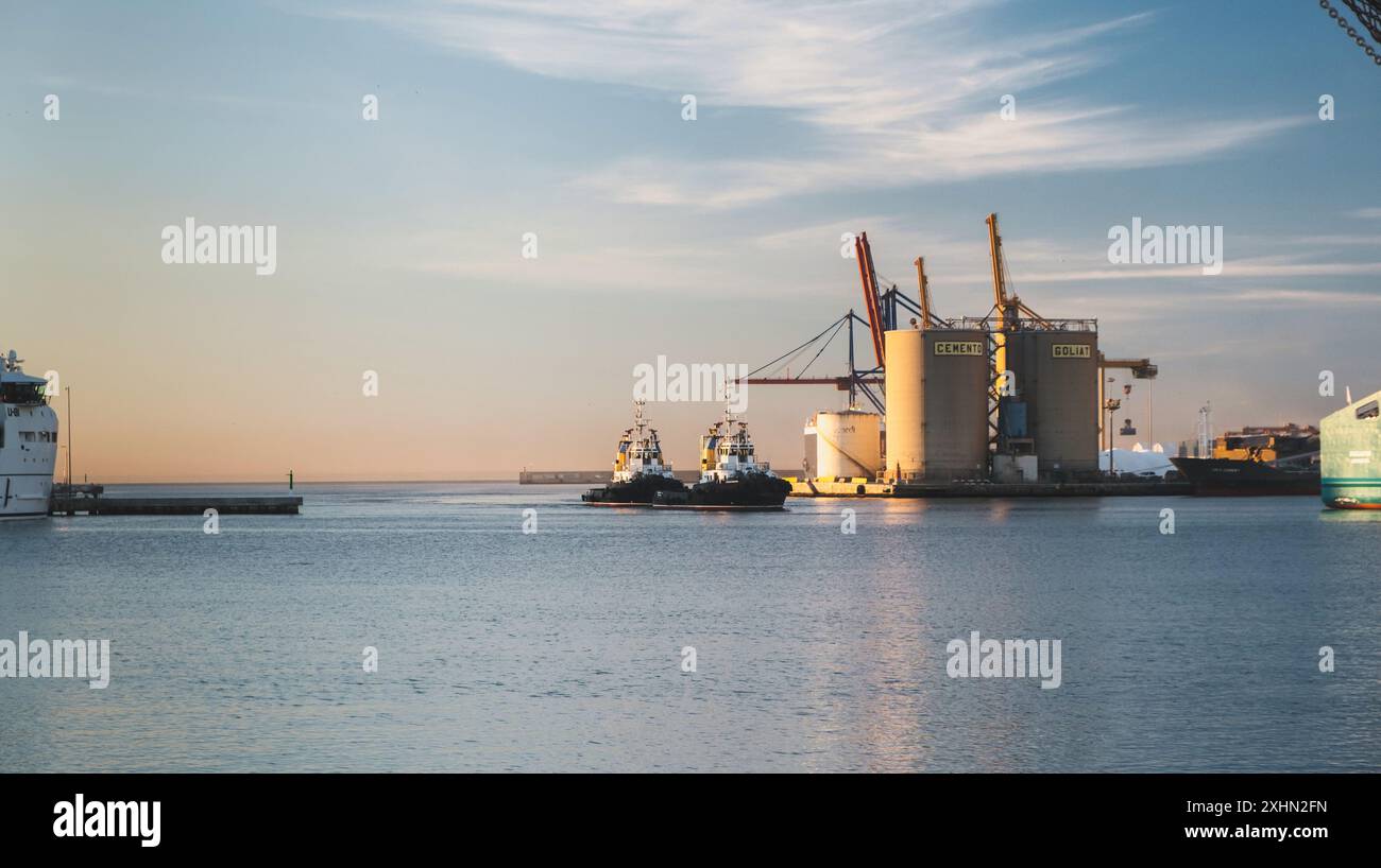Coucher de soleil scène de port maritime avec des navires de charge, remorqueurs, grues, et l'eau réfléchissante sereine au milieu des bâtiments industriels. Paysage maritime calme et fonctionnel Banque D'Images