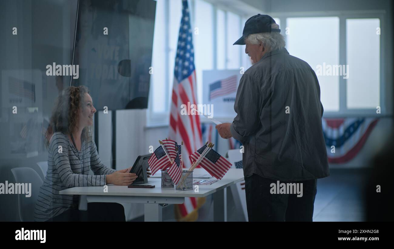 Citoyen américain mature caucasien se tient près de la table d'inscription au bureau de vote. Un officier de vote féminin parle à un homme, utilise une tablette et donne un bulletin de vote pour voter. Jour des élections aux États-Unis. Banque D'Images