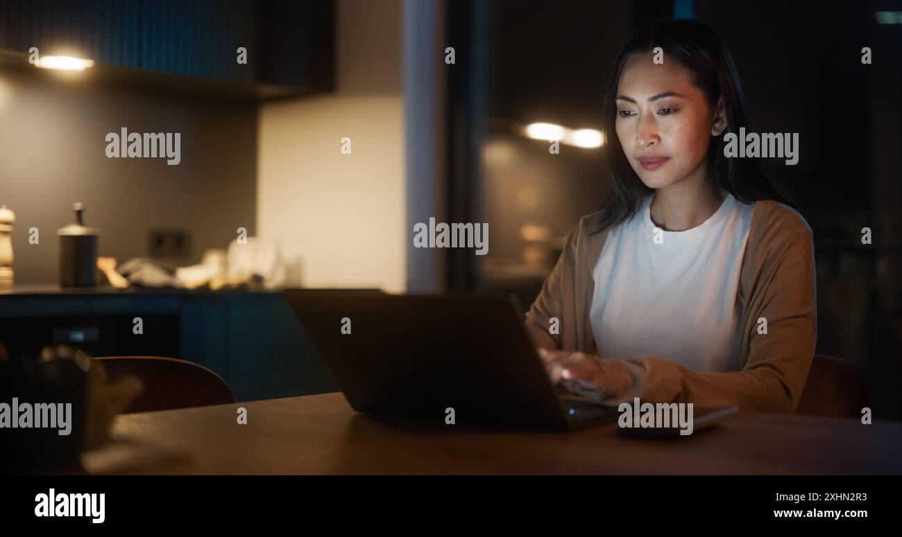 Belle femme asiatique authentique assise à une table dans une cuisine confortable et tapant sur ordinateur portable à la maison la nuit. Femme souriant et discutant sur les médias sociaux, faisant des achats en ligne, vérifiant les nouvelles. Banque D'Images