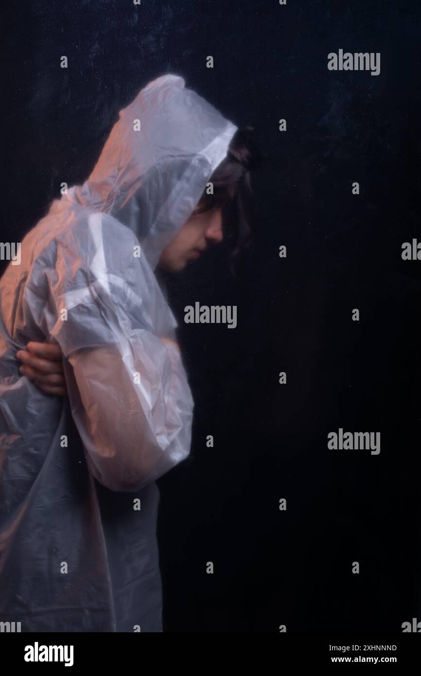 Jeune homme portant un imperméable en plastique transparent derrière le verre. Face cachée. Portrait de studio. Isolé sur fond sombre. Banque D'Images