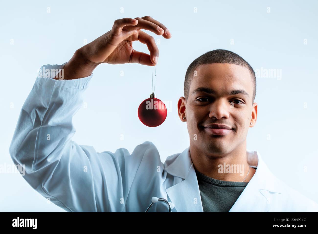 Docteur dans une blouse de laboratoire blanche, souriant et tenant un ornement de Noël rouge. En tant que médecin militaire, il cherche à améliorer la santé de ses patients pendant le fe Banque D'Images