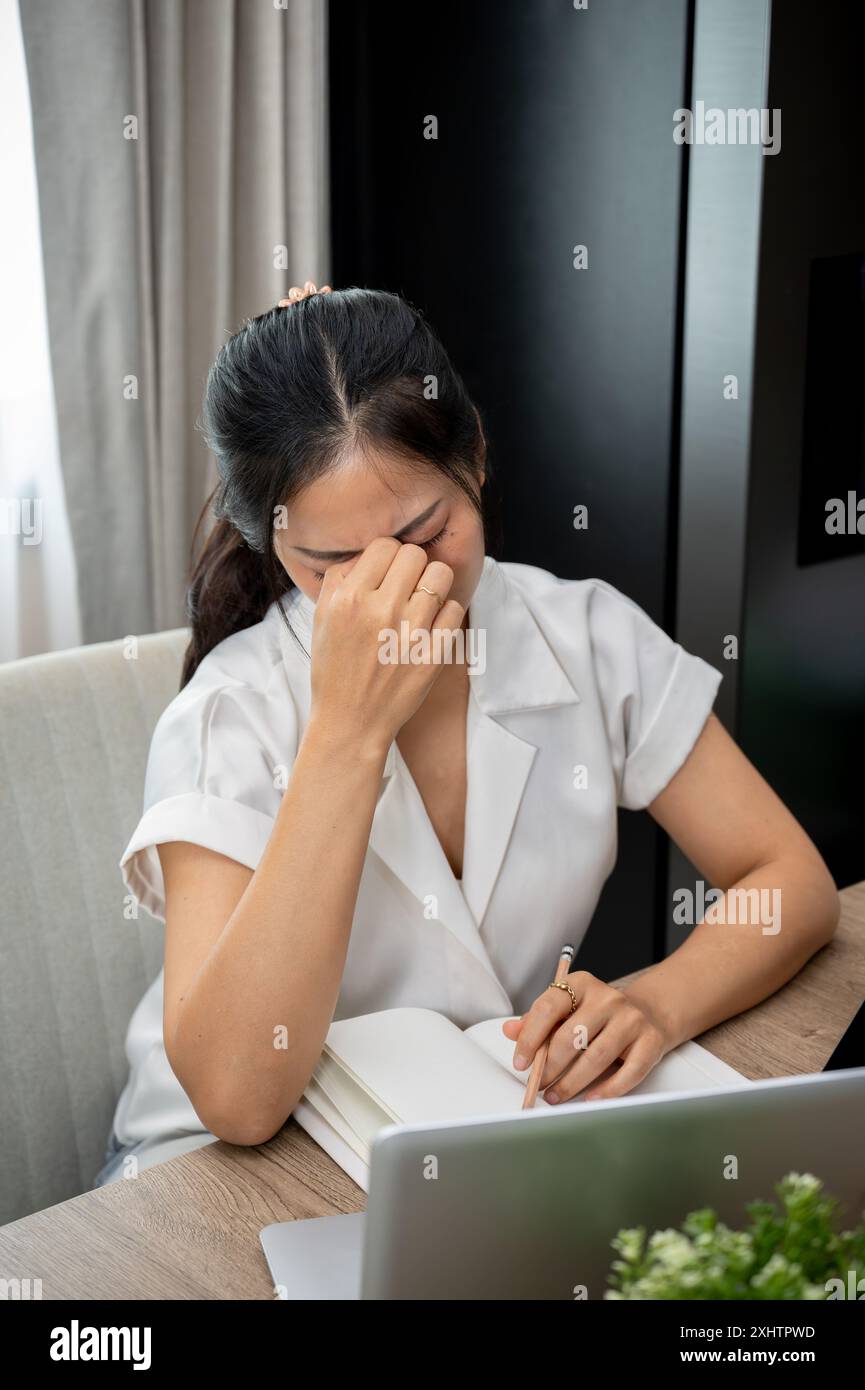Une femme asiatique stressée et frustrée souffre de maux de tête, réfléchit à des problèmes, s'inquiète de son travail, touche son visage, est assise à son bureau Banque D'Images