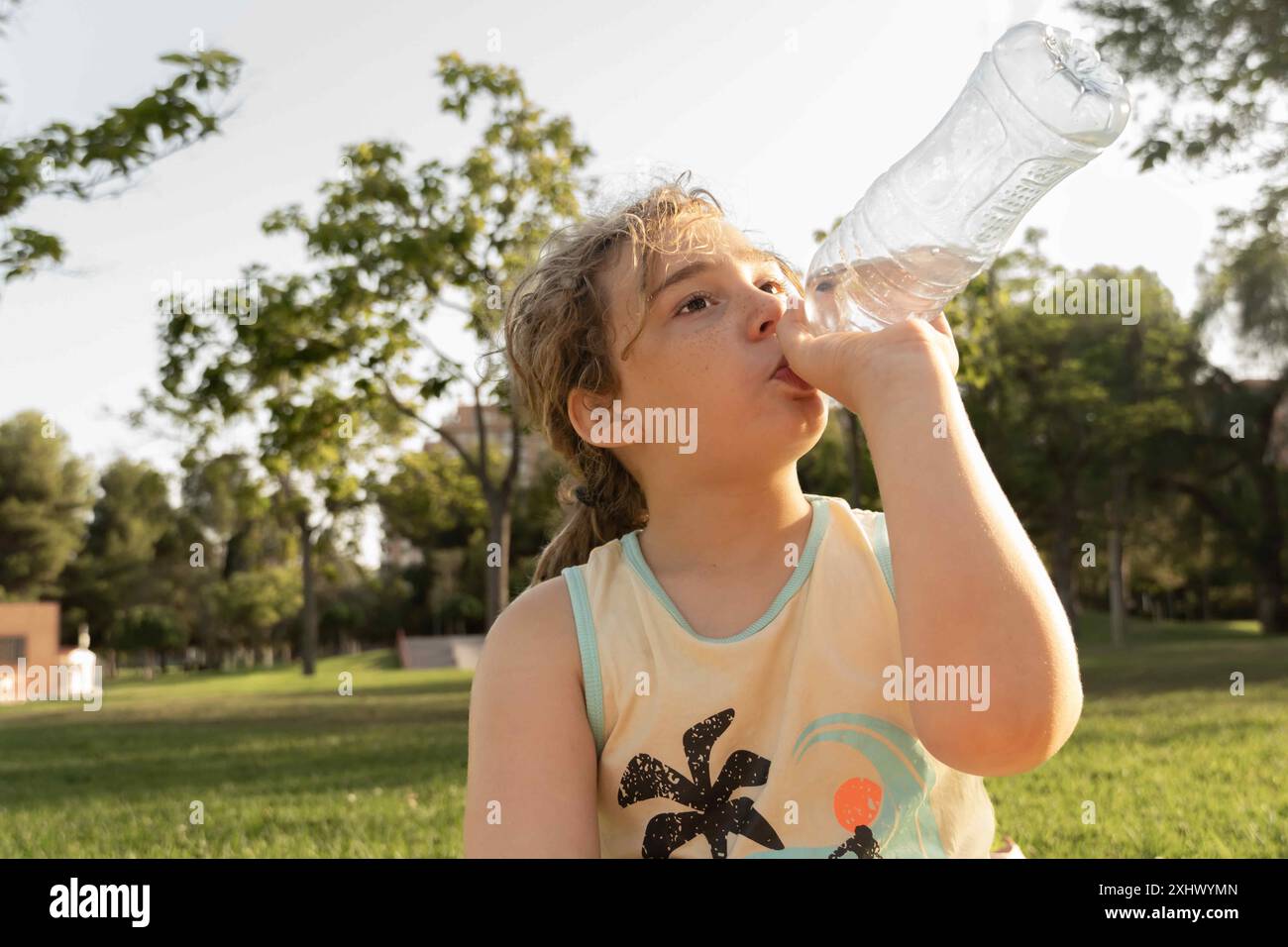 Gros plan d'un garçon aux cheveux clairs, buvant de l'eau pour se rafraîchir dans un parc ensoleillé. Journée chaude, avec la lumière du soleil et un environnement naturel, capturant un mome Banque D'Images