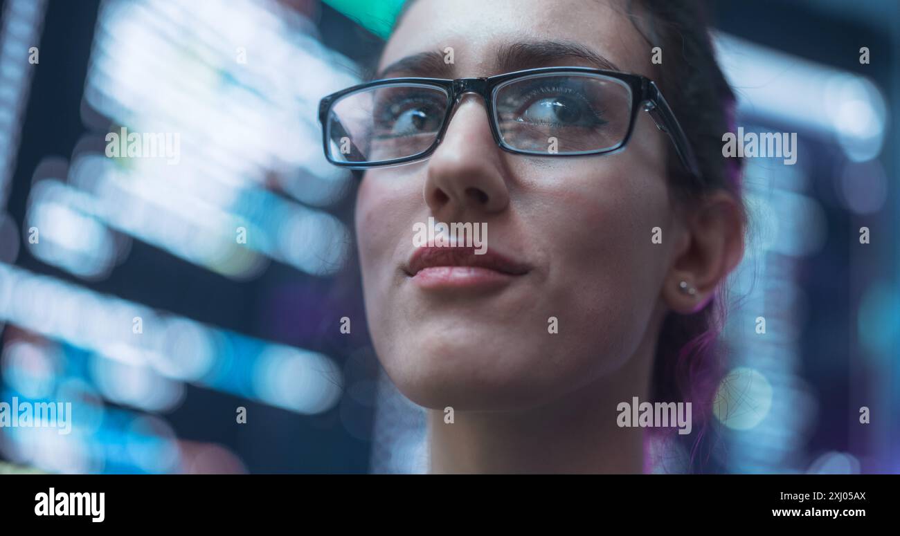 Gros plan Portrait d'une femme souriante regardant autour, lignes de langage de code reflétées sur ses lunettes à partir des grands écrans qui l'entourent. Programmeur professionnel femme développant des logiciels, codage Banque D'Images