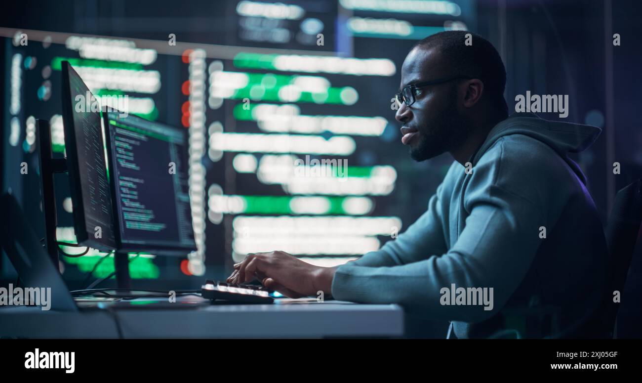 Programmeur masculin noir focalisé travaillant dans la salle de surveillance, entouré de grands écrans affichant des lignes de code de langage de programmation. Portrait de l'homme créant logiciel. Concept de codage futuriste. Banque D'Images
