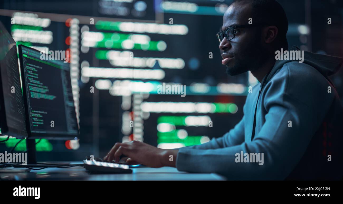 Programmeur masculin noir focalisé travaillant dans une salle de contrôle de surveillance, entouré de grands écrans affichant des lignes de code de langage de programmation. Portrait d'un homme créant un logiciel et un codage Banque D'Images