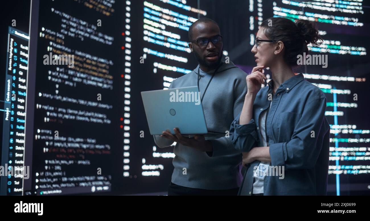 Portrait de deux développeurs divers utilisant un ordinateur portable, discutant des lignes de code qui apparaissent sur les grands écrans qui les entourent. Programmeurs masculins et féminins créer un logiciel ensemble et corriger des bugs Banque D'Images