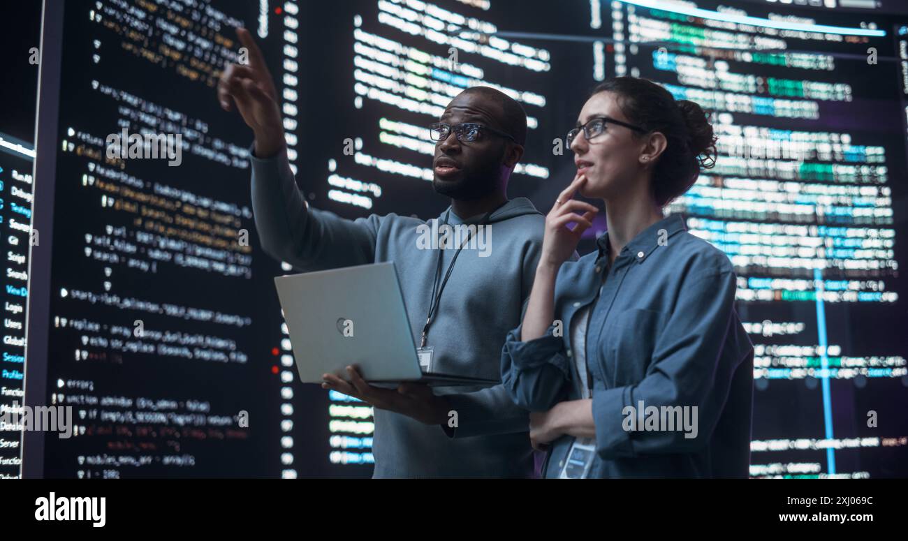 Portrait de deux développeurs divers utilisant un ordinateur portable, discutant des lignes de code qui apparaissent sur les grands écrans qui les entourent. Programmeurs masculins et féminins créant des logiciels ensemble et corrigeant des bugs Banque D'Images