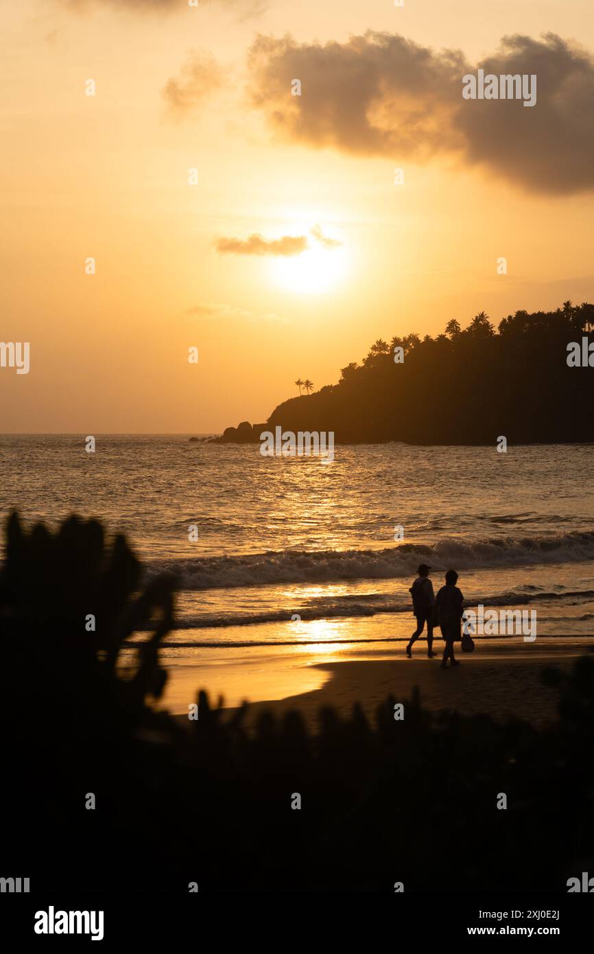 Silhouette de deux personnes marchant le long de la plage pendant l'heure dorée. Le soleil se couche et les vagues s'écrasent doucement sur le rivage. Banque D'Images