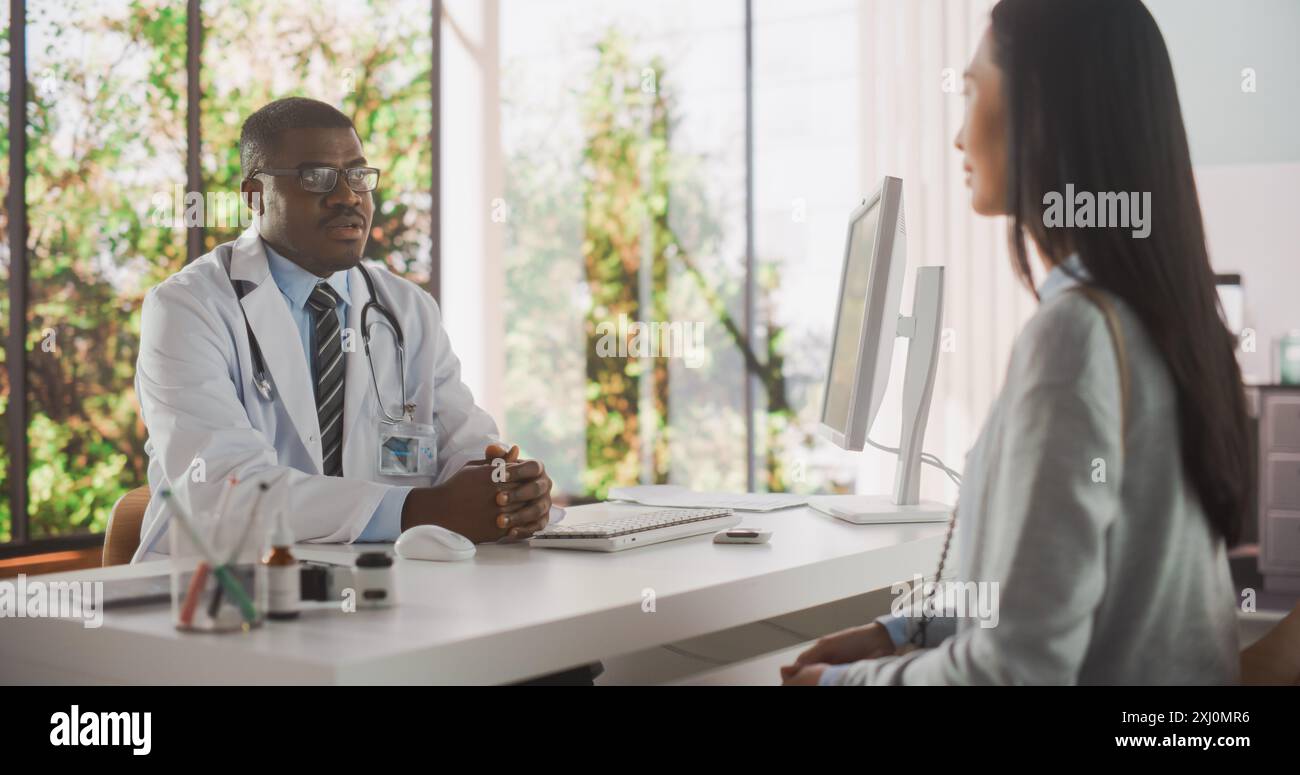 Handsome Young African Doctor a une conversation avec une patiente asiatique multiethnique, parlant avec elle lors d'une consultation dans une clinique de santé. Médecin en White Coat travaillant à l'hôpital moderne Banque D'Images