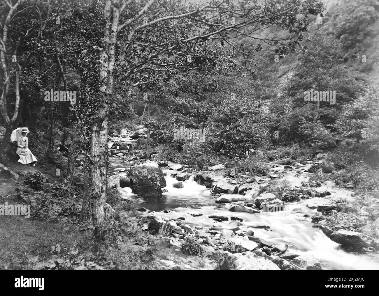 Paysage de la vallée du Lyn. Une dame avec un parasol assis à côté de la rivière Lyn. Lynmouth, Devon. Cette photographie est tirée d'un original édouardien, vers 1910. L'original faisait partie d'un album de 150 photographies d'albumen, de qualité variable, dont beaucoup j'ai photographié. La collection comprenait des images en particulier de l'île de Man et du comté anglais du Devonshire. Des annotations ont été incluses dans l'album mais, malheureusement, il n'y avait pas de dates précises. Les photos originales étaient en moyenne 6x4 ½ pouces. Banque D'Images