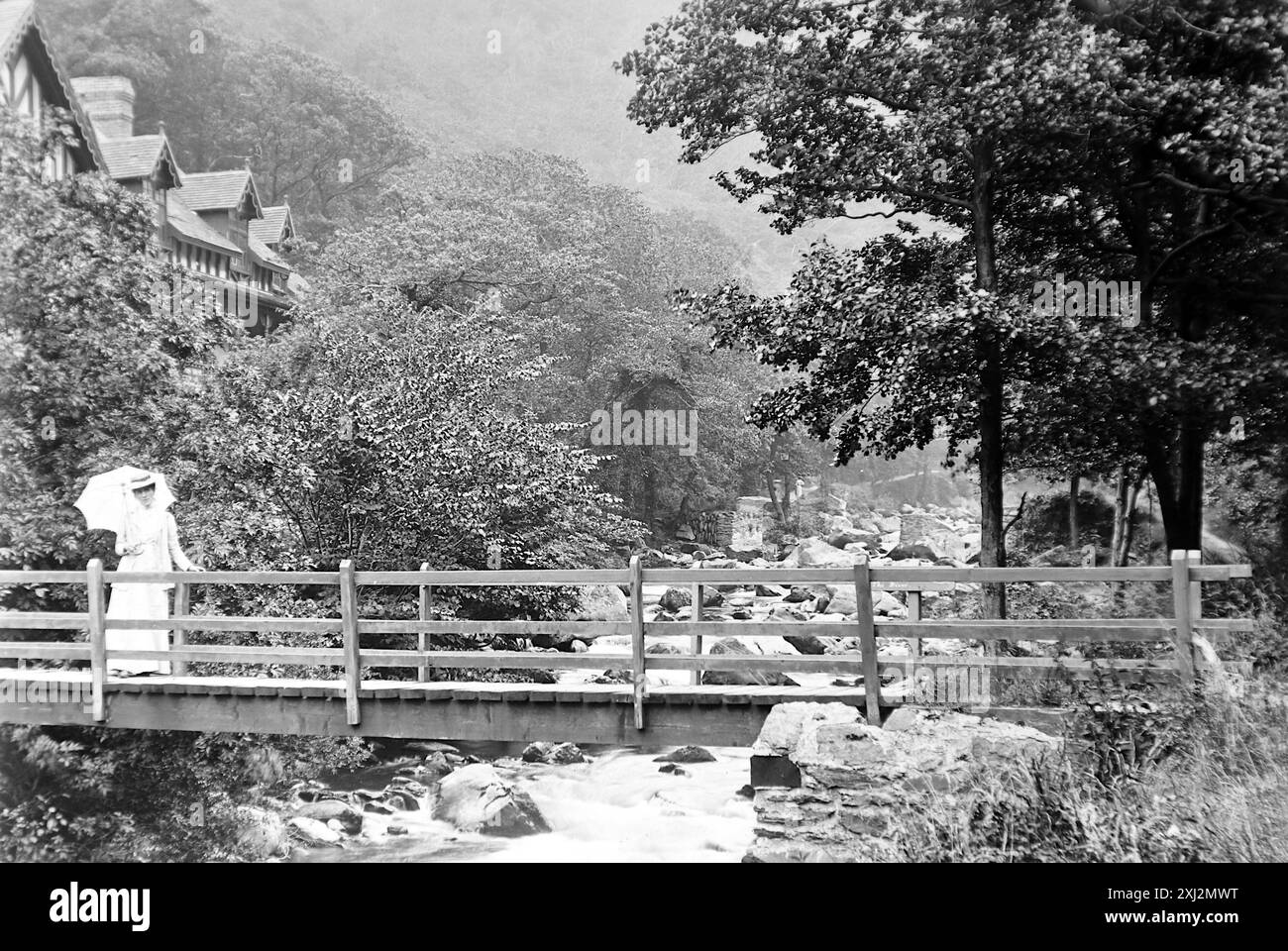 Lyn Valley, une passerelle sur la rivière Lyn avec une dame tenant un parasol. Lynmouth, Devon. Cette photographie est tirée d'un original édouardien, vers 1910. L'original faisait partie d'un album de 150 photographies d'albumen, de qualité variable, dont beaucoup j'ai photographié. La collection comprenait des images en particulier de l'île de Man et du comté anglais du Devonshire. Des annotations ont été incluses dans l'album mais, malheureusement, il n'y avait pas de dates précises. Les photos originales étaient en moyenne 6x4 ½ pouces. Banque D'Images