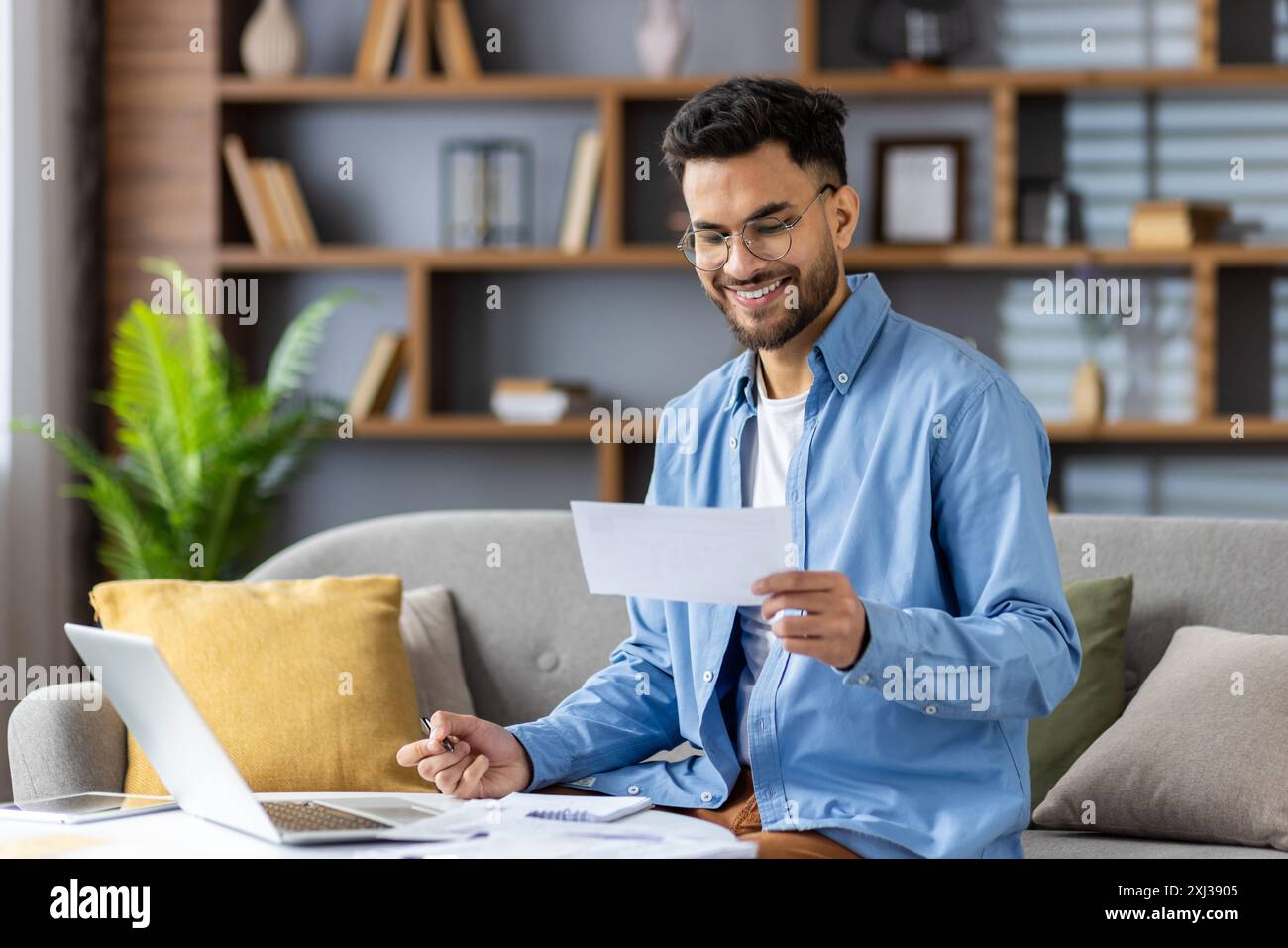 Jeune homme indien en lunettes et chemise bleue assis sur le canapé à la maison et travaillant à distance, vérifiant les factures et le budget du ménage. Banque D'Images
