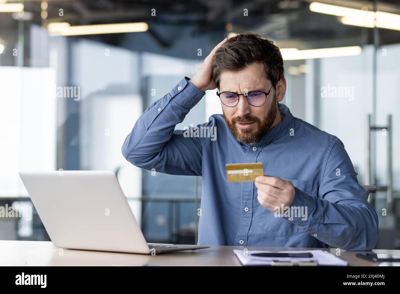 Jeune homme inquiet assis au bureau dans le bureau avec un ordinateur portable, les mains sur la tête et regardant la carte de crédit en état de choc. Banque D'Images
