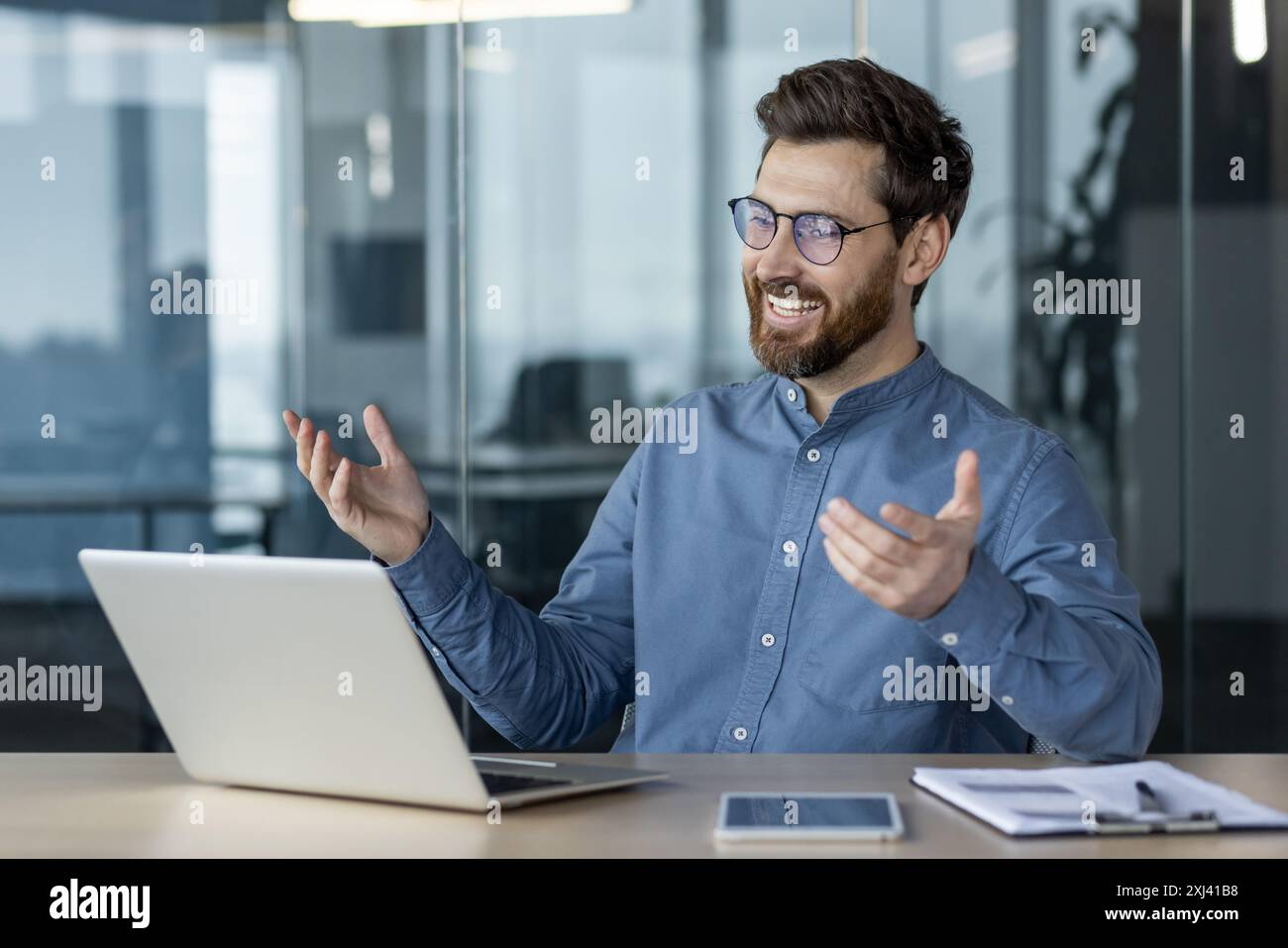 Souriant jeune homme d'affaires assis dans le bureau au bureau et parlant sur un appel vidéo sur l'ordinateur portable, faisant des gestes avec ses mains. Banque D'Images