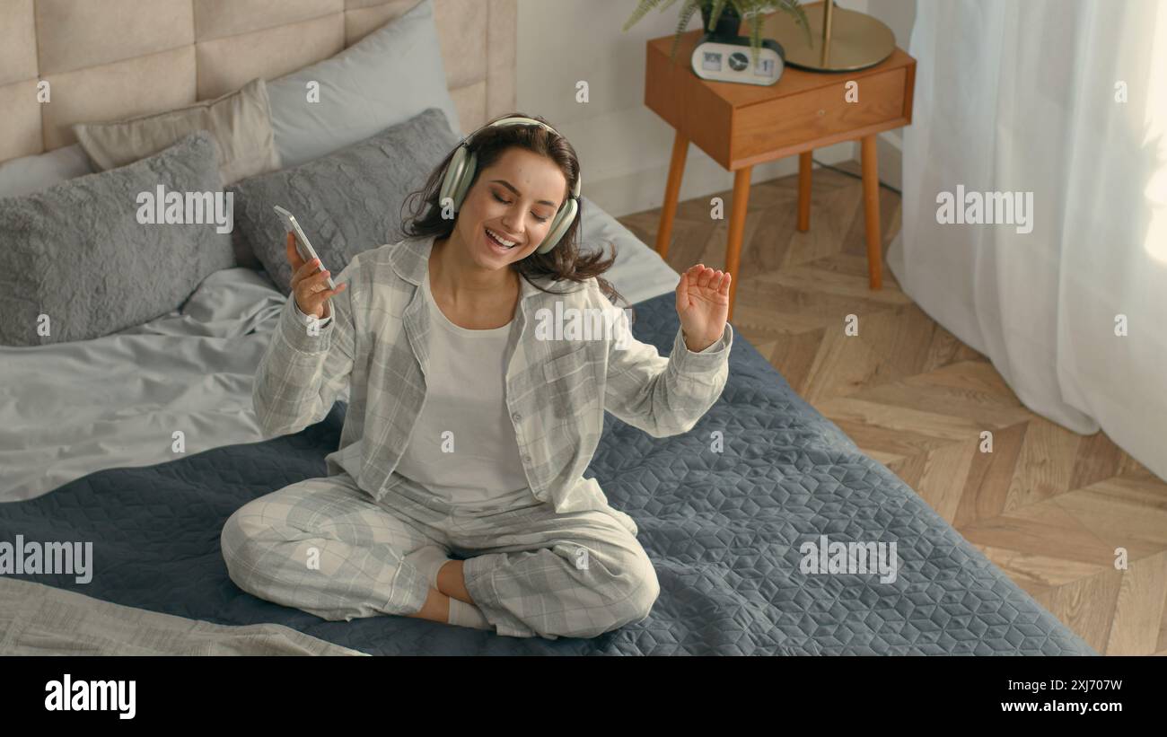 Vue de dessus heureuse femme caucasienne assise lit écouteurs smartphone téléphone mobile gadget technologie électronique écouter de la musique chantant fun énergique Banque D'Images