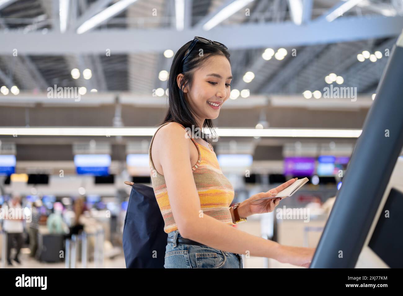 Une belle et positive passager touristique asiatique avec un passeport dans la main utilise un kiosque d'enregistrement libre-service dans le terminal de l'aéroport. peop Banque D'Images