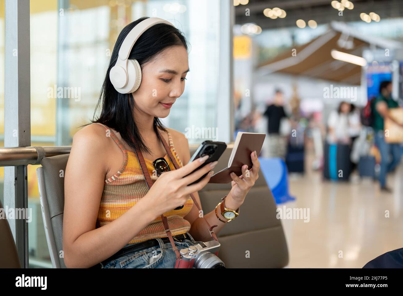 Une belle jeune touriste asiatique tient son smartphone et son passeport, vérifiant ses informations de vol sur son téléphone tout en étant assise à une attente Banque D'Images