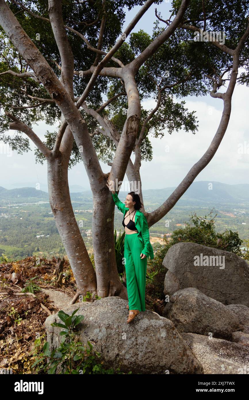 Une femme dans une tenue verte s'appuie contre un grand arbre sur une colline surplombant un paysage verdoyant. Banque D'Images