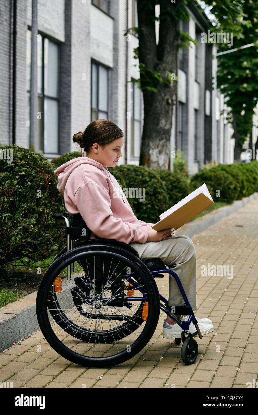 Une jeune femme en sweat à capuche rose est assise dans un fauteuil roulant lisant un livre sur un chemin de briques à l'extérieur d'un bâtiment. Banque D'Images