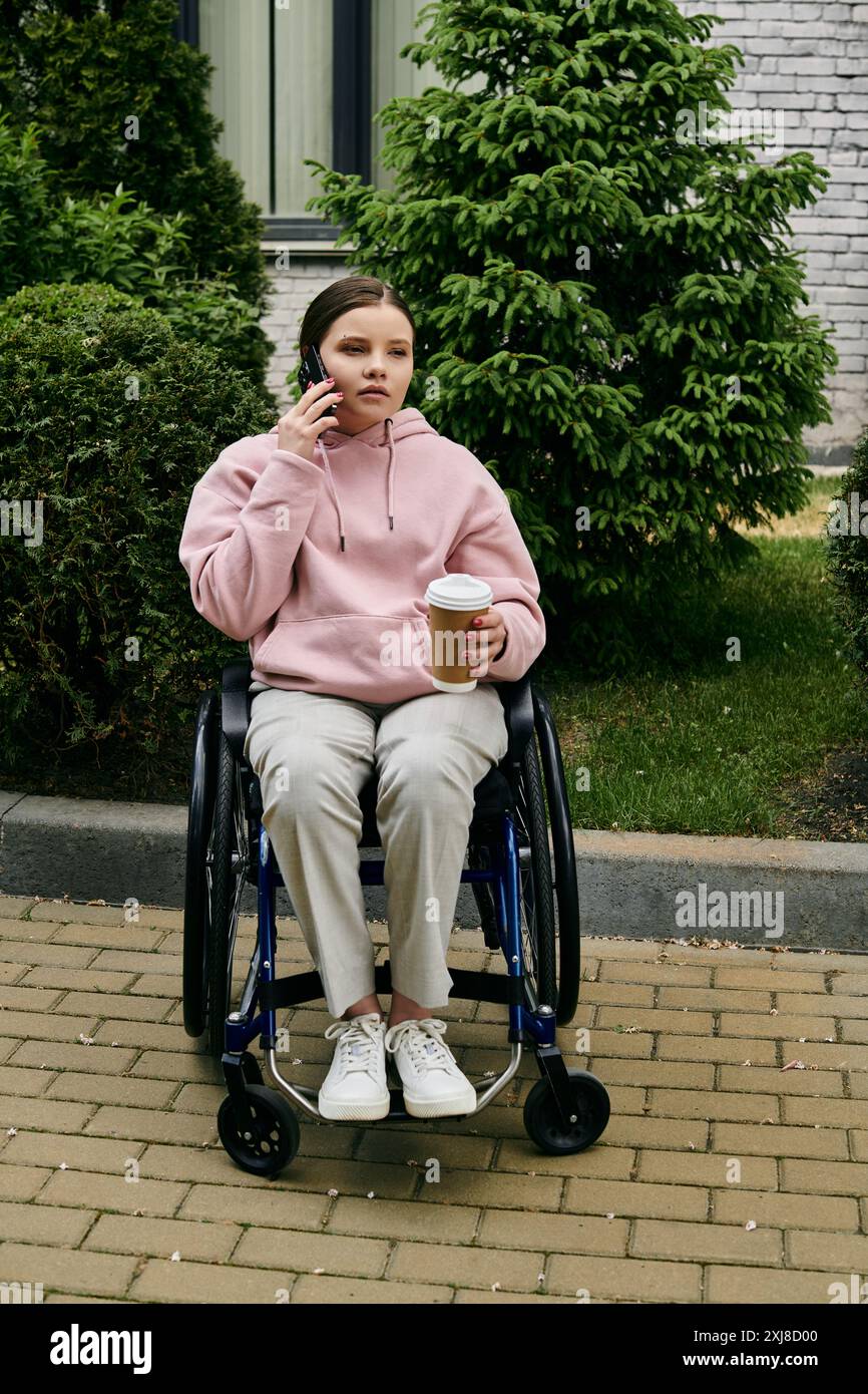 Une jeune femme en sweat à capuche rose est assise dans un fauteuil roulant, profitant d'un café et d'un appel téléphonique par une journée ensoleillée. Banque D'Images